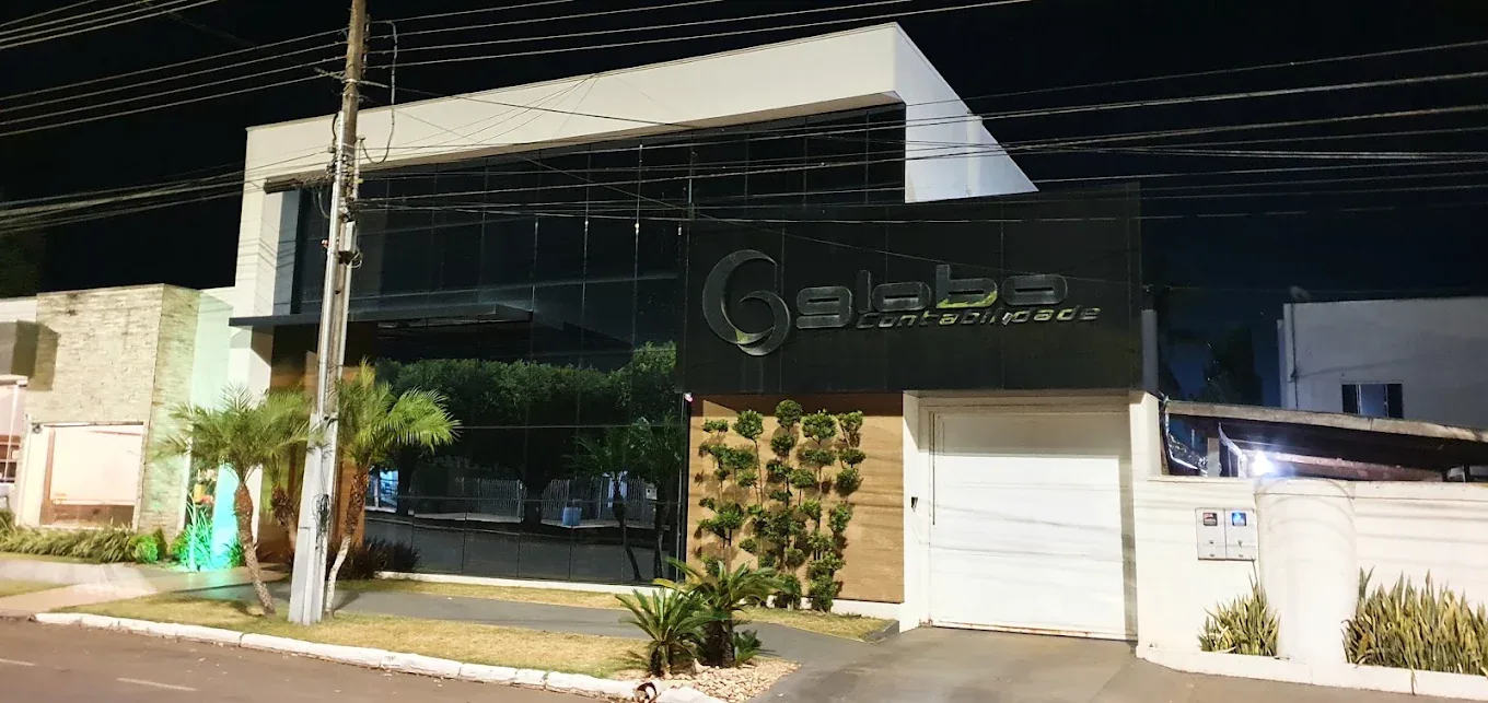 Fachada do escritório da Globo Contabilidade à noite: prédio de dois pavimentos revestido em vidro escuro, com o letreiro iluminado da empresa e jardim vertical na entrada.
